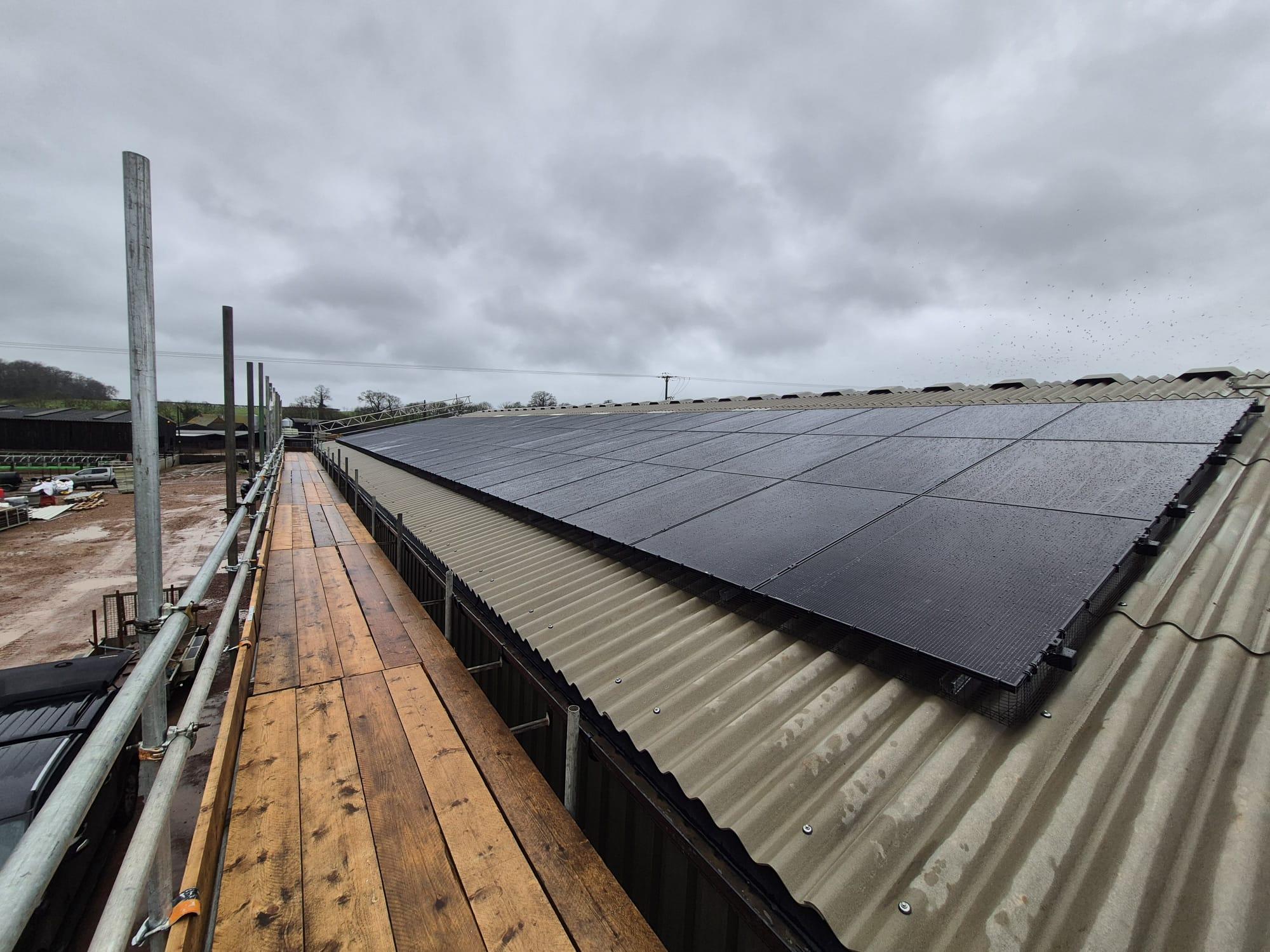 A long row of black solar panels on a fibre cement barn roof at Underhill Farm, seen from the scaffold walkway.